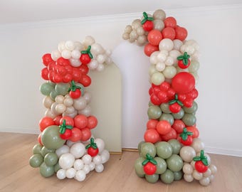 May include: Two balloon arches, one on the left and one on the right, are decorated with red, green, and cream-colored balloons. Each arch is adorned with red balloon "tomatoes" with green leaf accents, creating a festive and playful look. The arches are set against a white wall.