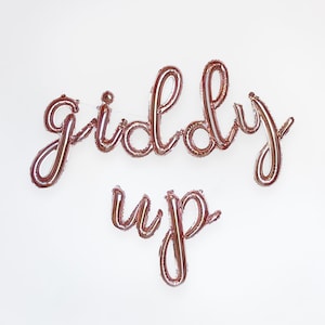 May include: Rose gold foil balloon letters spelling "giddy up" in a cursive font.