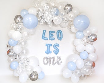May include: A balloon arch in shades of light blue, white, and silver, featuring clear balloons with confetti, a large silver snowflake foil balloon, and blue and silver foil letter balloons that spell out the text LEO IS ONE.