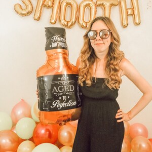 May include: A woman wearing sunglasses and a black dress holds a gold balloon shaped like a bottle of whisky. The balloon has a black label that says "Aged to Perfection". The background is decorated with gold balloons that spell out "SMOOTH" and colourful balloons.
