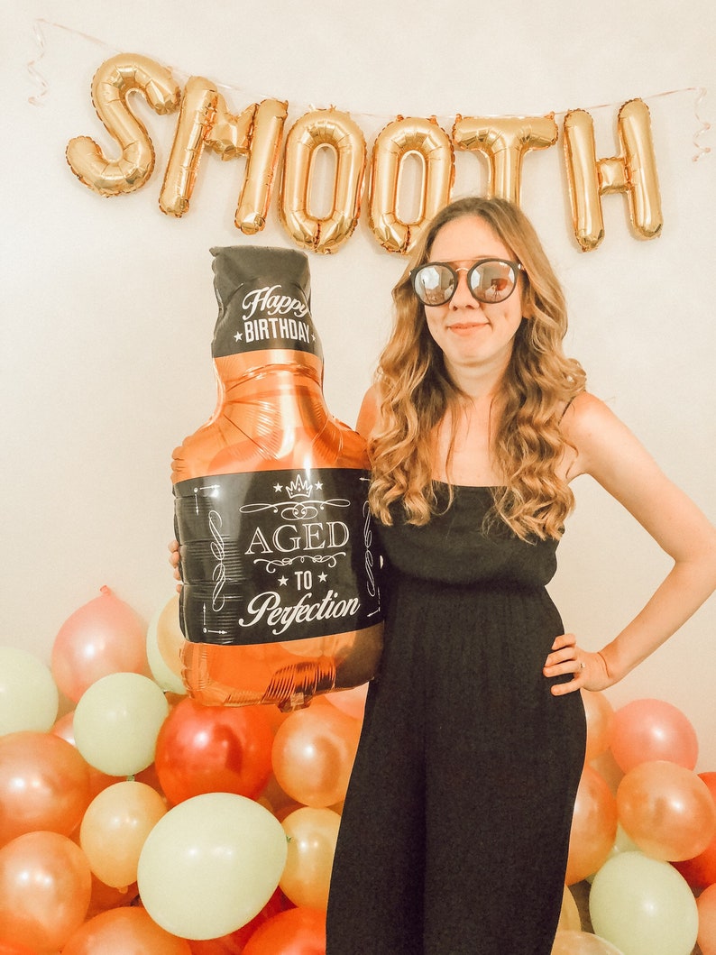 May include: A woman wearing sunglasses and a black dress holds a gold balloon shaped like a bottle of whisky. The balloon has a black label that says "Aged to Perfection". The background is decorated with gold balloons that spell out "SMOOTH" and colourful balloons.
