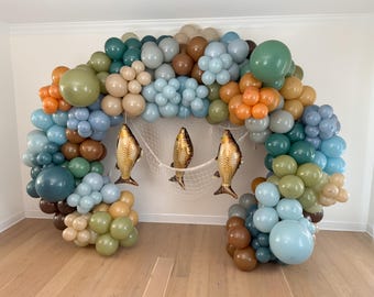 Puede incluir: Un arco de globos en tonos azules, verdes, marrones y naranjas, decorado con tres peces dorados y una red blanca. El arco está colocado contra una pared blanca y un suelo de madera, creando un ambiente festivo.