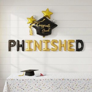 May include: A graduation-themed balloon arrangement. A black mortarboard balloon with gold text reading "Congrats Grad" is surrounded by gold and silver star balloons. Below, the word "PHINISHED" is spelled out in gold and black balloons. A graduation cap and diploma are on a table.