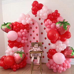 May include: A strawberry-themed party setup with a pink backdrop adorned with strawberry illustrations. The arch is decorated with red, pink, and white balloons, and strawberry-shaped foil balloons. A child sits in a wooden high chair.