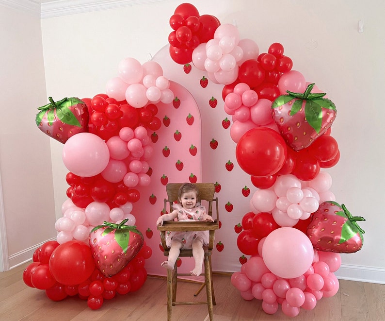 May include: A strawberry-themed party setup with a pink backdrop adorned with strawberry illustrations. The arch is decorated with red, pink, and white balloons, and strawberry-shaped foil balloons. A child sits in a wooden high chair.
