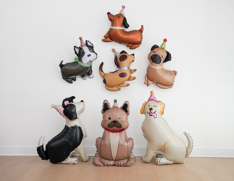 May include: A collection of dog-shaped balloons in various colors, each wearing a party hat. The balloons are arranged against a white wall and on a wooden floor, creating a festive and playful atmosphere, perfect for a birthday celebration.