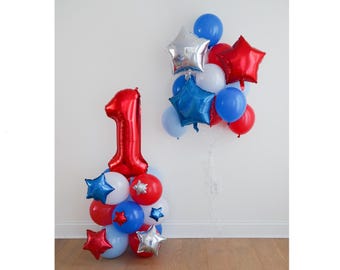 May include: A balloon arrangement featuring a large red foil number one balloon atop a cluster of red, white, and blue latex balloons and small star-shaped foil balloons, alongside a floating bouquet of red, white, and blue balloons.
