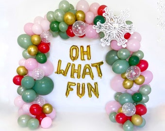 May include: A balloon arch featuring sage green, light pink, red, and gold balloons, along with clear balloons containing white snowflake confetti. A large white snowflake cutout and gold foil balloons spelling out OH WHAT FUN are included.