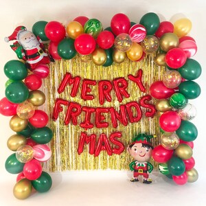 May include: A festive balloon arch frame featuring red, dark green, and gold balloons, including clear balloons filled with gold confetti. A foil Santa Claus balloon is on the top left, a foil elf balloon is on the bottom right, and red foil letters spell MERRY FRIENDS MAS over a gold tinsel curtain.