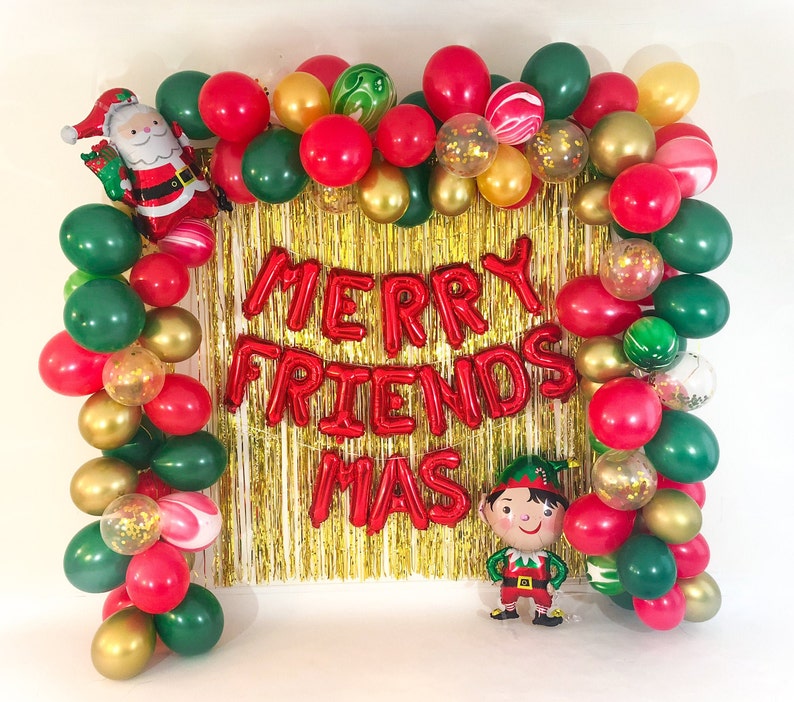 May include: A festive balloon arch frame featuring red, dark green, and gold balloons, including clear balloons filled with gold confetti. A foil Santa Claus balloon is on the top left, a foil elf balloon is on the bottom right, and red foil letters spell MERRY FRIENDS MAS over a gold tinsel curtain.