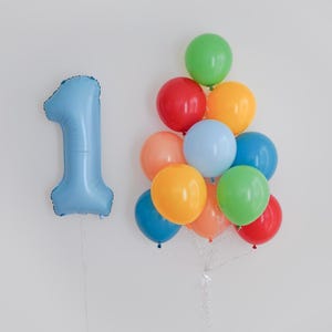 May include: A light blue foil balloon shaped like the number one positioned next to a cluster of eleven round latex balloons in various colors including red, orange, yellow, green, and blue, all tied together with white ribbons.