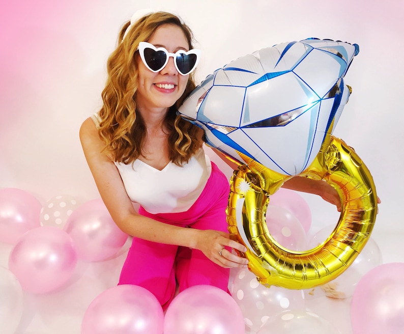 May include: A woman wearing heart-shaped sunglasses holds a large gold and silver ring balloon. The balloon is shaped like a diamond ring. The woman is surrounded by pink and white polka dot balloons.