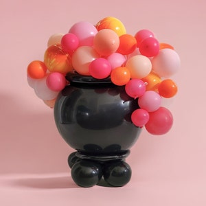 May include: A black cauldron with a round base and three legs is decorated with a cluster of pink, orange, and yellow balloons.