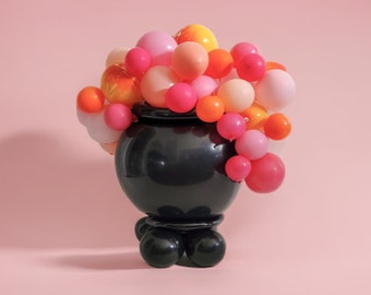 May include: A black cauldron with a round base and three legs is decorated with a cluster of pink, orange, and yellow balloons.
