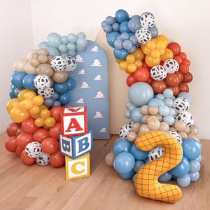 Puede incluir: Un arco de globos colorido con globos azules, marrones, amarillos y rojos, acentuado con globos con estampado de d&aacute;lmata. Bloques de letras de madera deletrean "ABC". Tambi&eacute;n hay un gran globo amarillo con el n&uacute;mero "2". El tel&oacute;n de fondo es un panel azul con nubes blancas.