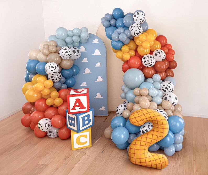 Puede incluir: Un arco de globos colorido con globos azules, marrones, amarillos y rojos, acentuado con globos con estampado de d&aacute;lmata. Bloques de letras de madera deletrean "ABC". Tambi&eacute;n hay un gran globo amarillo con el n&uacute;mero "2". El tel&oacute;n de fondo es un panel azul con nubes blancas.