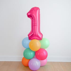 May include: A large, metallic hot pink foil balloon shaped like the number one, positioned atop a cluster of smaller, round latex balloons in various pastel and bright colors including light blue, yellow, green, orange, pink, and purple.