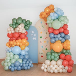 May include: Two arched backdrops decorated with multi-colored balloon garlands in shades of sage green, cream, orange, red, and blue. The blue arch features the text "FUN TO BE ONE" in white, while the cream arch displays colorful confetti patterns.