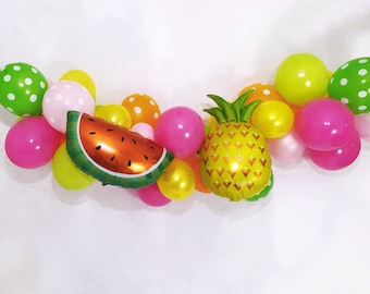 Op de afbeelding: Een ballonslinger met een mix van ronde ballonnen in tinten roze, geel, oranje, limoengroen en witte stippen, geaccentueerd door twee folieballonnen in de vorm van een watermeloenpartje en een hele ananas met groene bladeren.