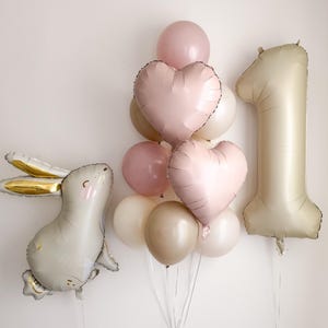 Puede incluir: Un conjunto de globos para una celebraci&oacute;n de primer cumplea&ntilde;os. Incluye un gran globo con el n&uacute;mero uno, un globo con forma de conejito y un grupo de globos en forma de coraz&oacute;n y redondos en tonos rosa, blanco y dorado.