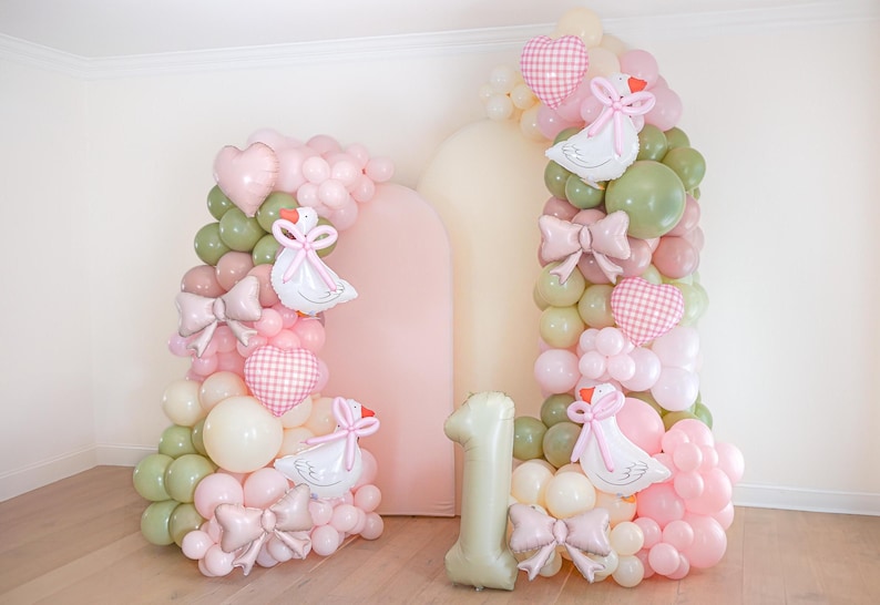 May include: A festive balloon display in the shape of the number one, with a backdrop. The balloons are in shades of pink, green, and white, with heart and bow accents, and swan-shaped balloons. A perfect decoration for a first birthday.