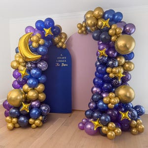 May include: Two balloon arches, one with a gold crescent moon and the other with a large gold sphere. The balloons are in shades of gold, blue, purple, and star-printed. A navy blue backdrop reads "A Night Under The Stars."