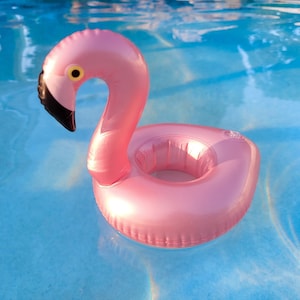 K&ouml;nnte beinhalten: Ein rosa aufblasbarer Flamingo-Pool-Schwimmer mit schwarzem Schnabel und gelbem Auge schwimmt in einem blauen Schwimmbecken. Der Flamingo hat eine kreisf&ouml;rmige &Ouml;ffnung f&uuml;r ein Getr&auml;nk oder eine Tasse.