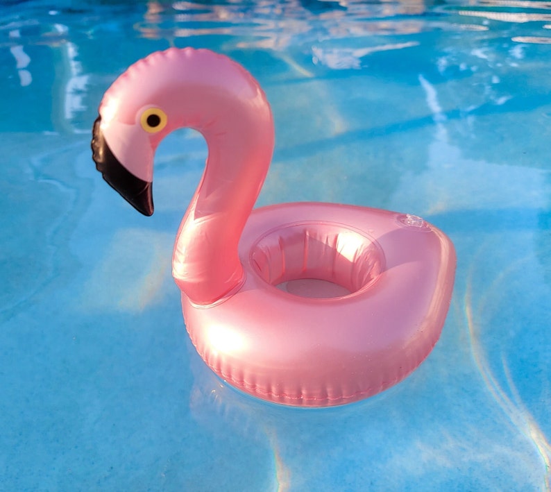 K&ouml;nnte beinhalten: Ein rosa aufblasbarer Flamingo-Pool-Schwimmer mit schwarzem Schnabel und gelbem Auge schwimmt in einem blauen Schwimmbecken. Der Flamingo hat eine kreisf&ouml;rmige &Ouml;ffnung f&uuml;r ein Getr&auml;nk oder eine Tasse.