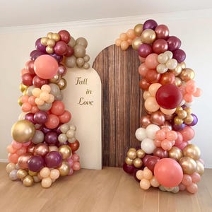 May include: A balloon arch decoration featuring a color palette of burgundy, peach, gold, and cream balloons. The arrangement frames two arched panels, one with a wood grain texture and one with the text Fall in Love printed in gold script.