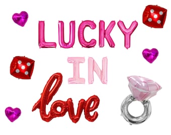 May include: A set of foil balloons featuring the text LUCKY IN LOVE in pink, light pink, and red. The set includes two red dice balloons with white dots, three small magenta heart balloons, and one large pink and silver diamond ring balloon.