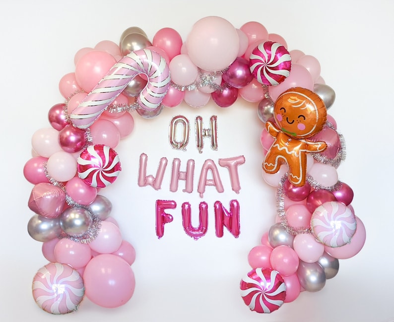 May include: Pink, silver, and brown balloon arch with candy cane, gingerbread man, and swirl designs. The arch spells out "Oh What Fun" in pink and silver letters.