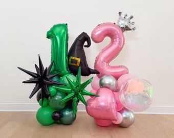 May include: A festive balloon arrangement featuring the numbers 1 and 2 in green and pink, with a witch hat and crown. The arrangement includes star-shaped and round balloons in various colors, perfect for a birthday celebration.