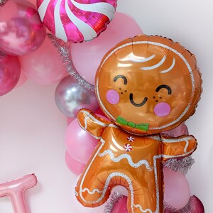 May include: A gingerbread man balloon with a smiling face and a green bow tie, surrounded by pink and silver balloons and tinsel.
