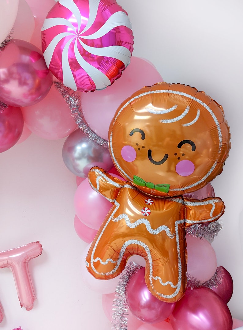 May include: A gingerbread man balloon with a smiling face and a green bow tie, surrounded by pink and silver balloons and tinsel.
