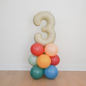 May include: A balloon arrangement featuring a large, beige number 3 balloon atop a cluster of colorful balloons. The balloons below are in shades of red, orange, green, and blue, creating a festive display. Perfect for a birthday celebration.