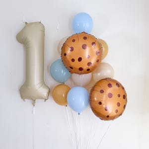 May include: A collection of balloons for a first birthday celebration. Includes a large gold number one balloon, cookie-shaped balloons, and smaller balloons in light blue, tan, and cream colors. The balloons are tied together with clear string.