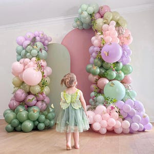 May include: A young child in a green dress with butterfly wings stands before a colorful balloon arch. The arch features pastel pink, green, and purple balloons, adorned with butterfly decorations. The scene evokes a whimsical, celebratory atmosphere.