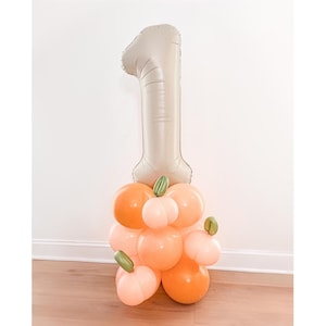 May include: A festive balloon arrangement for a first birthday celebration. A large, neutral-toned number one balloon sits atop a cluster of orange and peach-colored balloons, some adorned with green leaf accents. The balloons are arranged on a wooden floor.