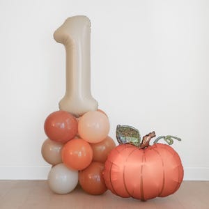 May include: A beige number one balloon sits atop a cluster of round balloons in shades of terracotta, peach, cream, and tan. Beside the balloon cluster is a large, round, metallic orange pumpkin-shaped balloon with a green stem accent.