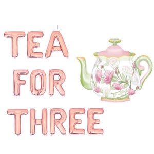 Puede incluir: Una tetera decorativa con un dise&ntilde;o floral y las palabras "TEA FOR THREE" en letras de globo de oro rosa. La tetera tiene detalles en rosa, verde y blanco.