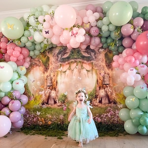 May include: A whimsical party backdrop featuring a green, pink, and white balloon arch with butterfly decorations. A young girl in a green dress stands in front of the arch, which is set against a backdrop of a forest scene.