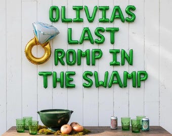 May include: Green foil letter balloons spell OLIVIAS LAST ROMP IN THE SWAMP above a table with a gold ring balloon, a dark green bowl, two onions, and several green glasses and beverage cans arranged on a wooden surface.