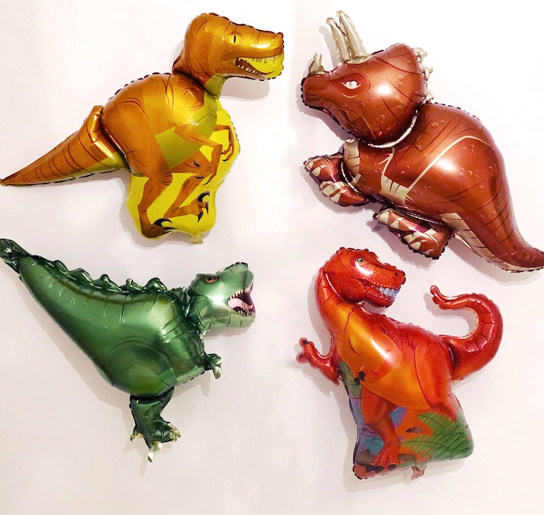 Puede incluir: Cuatro globos de aluminio con forma de dinosaurios. Un T-Rex amarillo y naranja, un triceratops marr&oacute;n, un T-Rex verde met&aacute;lico con la boca abierta y un T-Rex rojo y naranja con una cola curva y detalles de follaje verde.