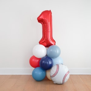 May include: A balloon arrangement featuring a large red foil number one balloon atop a cluster of smaller round balloons in shades of red, white, navy blue, and light blue, accompanied by a white round balloon printed with red baseball stitching.