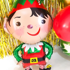 May include: A foil balloon shaped like an elf with a green hat featuring a red and white candy cane stripe, a red tunic with a yellow belt buckle, and black shoes with yellow accents. The elf has black hair, rosy cheeks, and a wide smile.