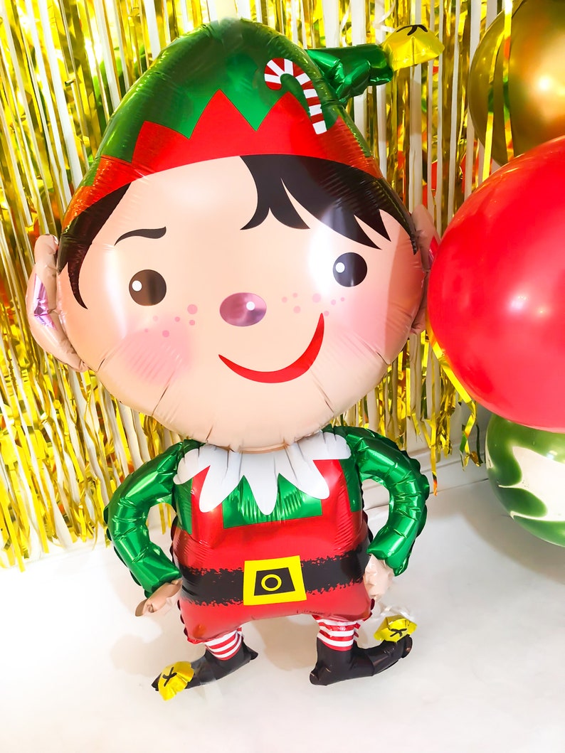 May include: A foil balloon shaped like an elf with a green hat featuring a red and white candy cane stripe, a red tunic with a yellow belt buckle, and black shoes with yellow accents. The elf has black hair, rosy cheeks, and a wide smile.