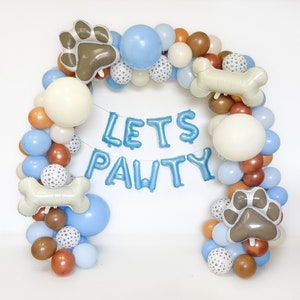 Puede incluir: Un arco de globos en tonos azules, blancos, marrones y naranjas, con globos con forma de huellas de patas y huesos. La frase "LETS PAWTY" est&aacute; escrita con globos de aluminio azules, creando una decoraci&oacute;n festiva para una fiesta tem&aacute;tica de mascotas.