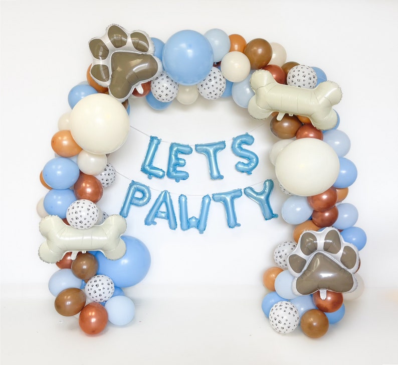 Puede incluir: Un arco de globos en tonos azules, blancos, marrones y naranjas, con globos con forma de huellas de patas y huesos. La frase "LETS PAWTY" est&aacute; escrita con globos de aluminio azules, creando una decoraci&oacute;n festiva para una fiesta tem&aacute;tica de mascotas.