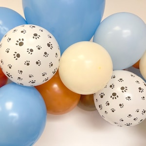 Puede incluir: Un conjunto de globos coloridos en tonos azul, marr&oacute;n y crema. Dos globos blancos est&aacute;n estampados con un patr&oacute;n de peque&ntilde;as huellas de patas negras. Los globos est&aacute;n dispuestos juntos, creando una exhibici&oacute;n festiva.