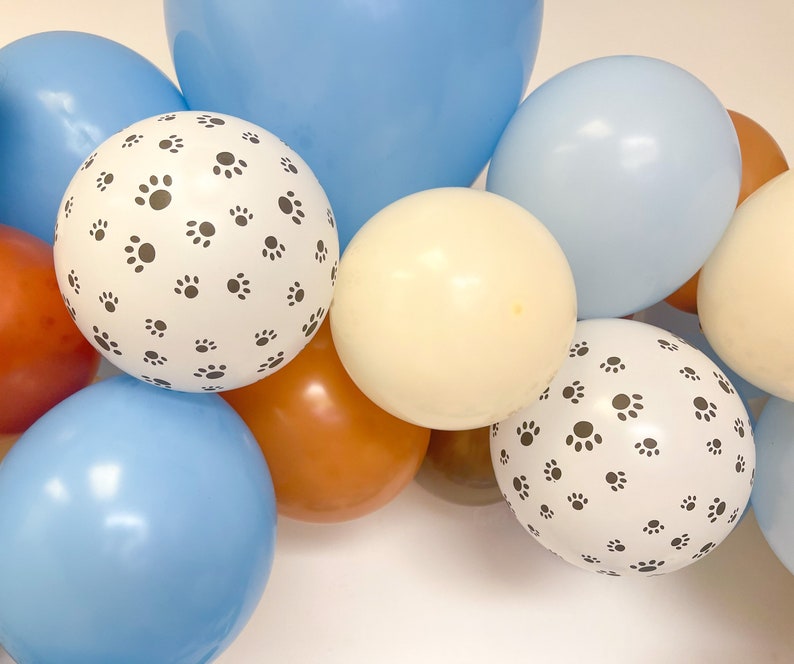 Puede incluir: Un conjunto de globos coloridos en tonos azul, marr&oacute;n y crema. Dos globos blancos est&aacute;n estampados con un patr&oacute;n de peque&ntilde;as huellas de patas negras. Los globos est&aacute;n dispuestos juntos, creando una exhibici&oacute;n festiva.
